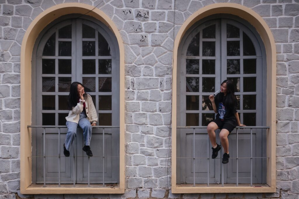 Two girls sitting in a window waving to each other.