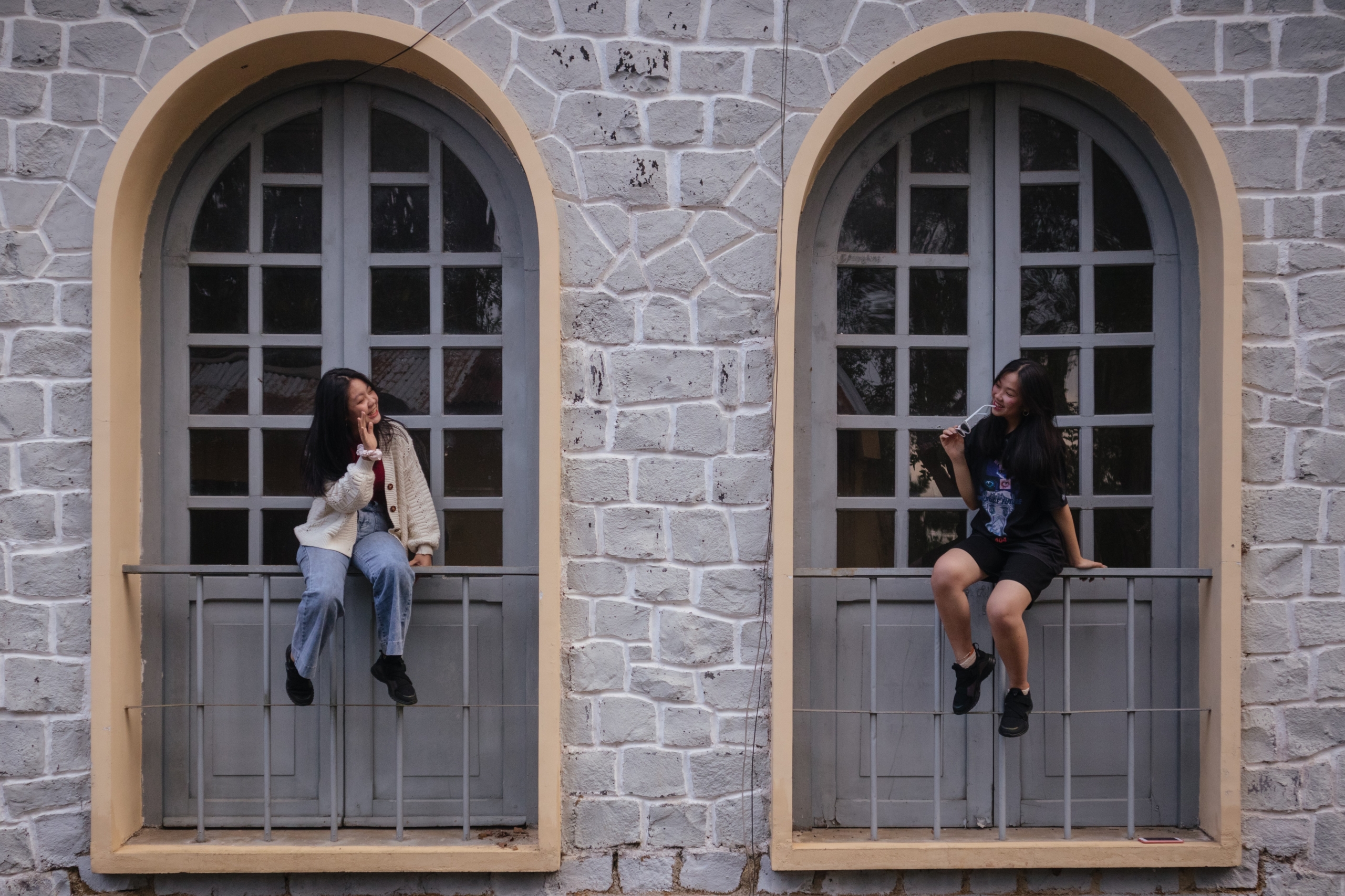 Two girls sitting in a window waving to each other.